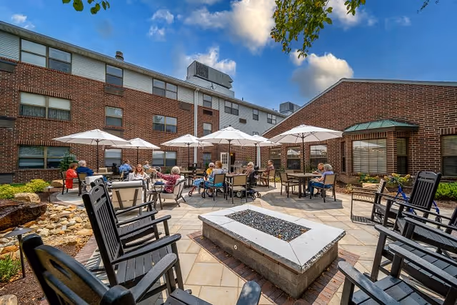 Outdoor patio area at Canton Regency with several elderly people sitting at tables under white umbrellas. The patio is surrounded by brick buildings and has black rocking chairs arranged around a rectangular fire pit. The sky is blue with some clouds.