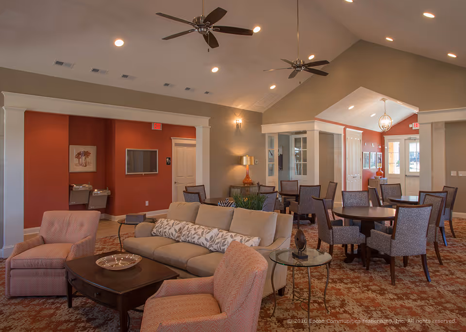 Spacious senior living community lounge with sofas, armchairs, dining tables, ceiling fans, and warm lighting.