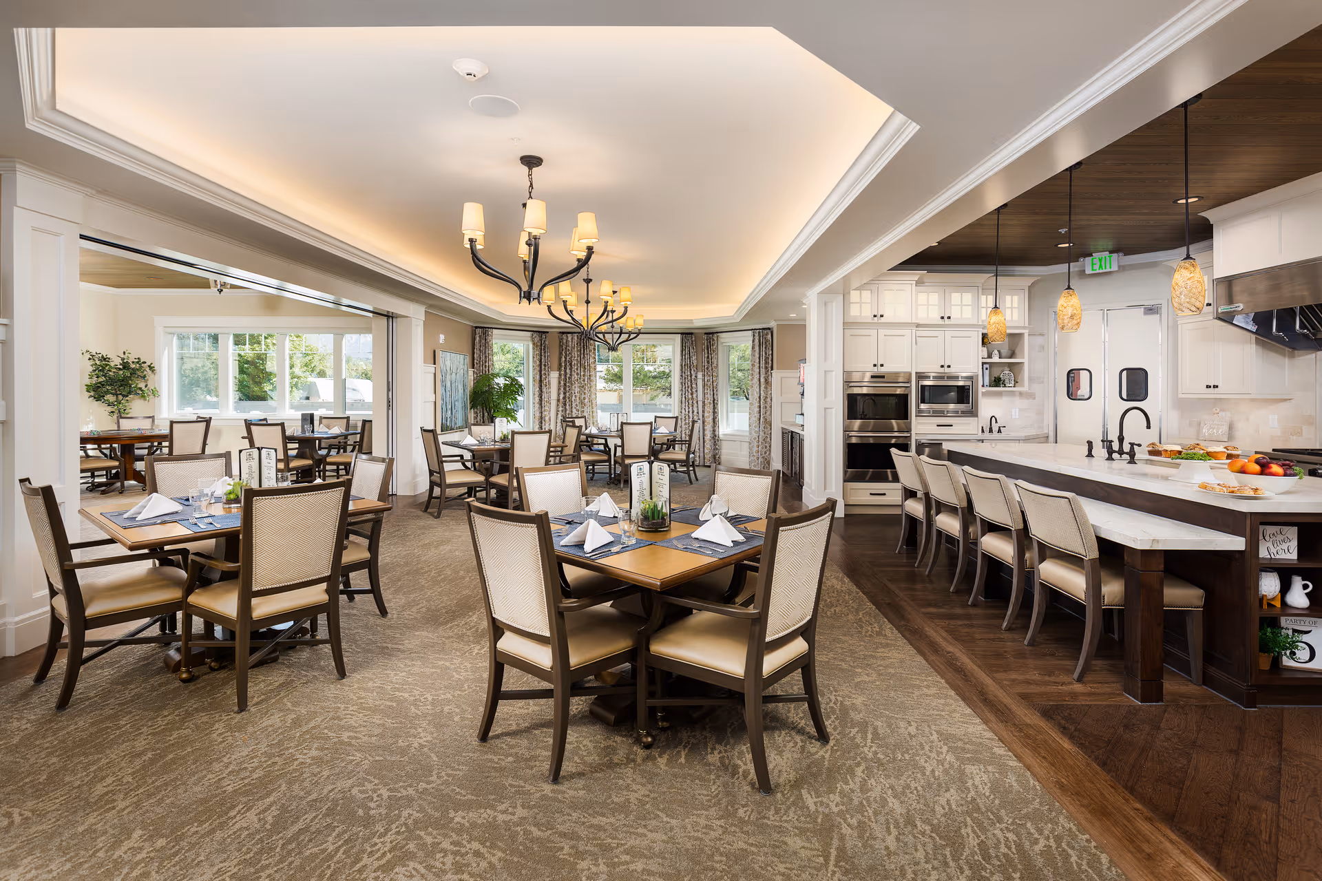 A spacious and well-lit dining area in a senior living facility featuring multiple wooden tables with chairs arranged neatly. Each table is set with placemats, napkins, and tableware. To the right, there is a modern kitchen area with a long counter and several high chairs. The room has large windows allowing natural light to fill the space, elegant chandeliers hanging from the ceiling, and a warm, inviting atmosphere.