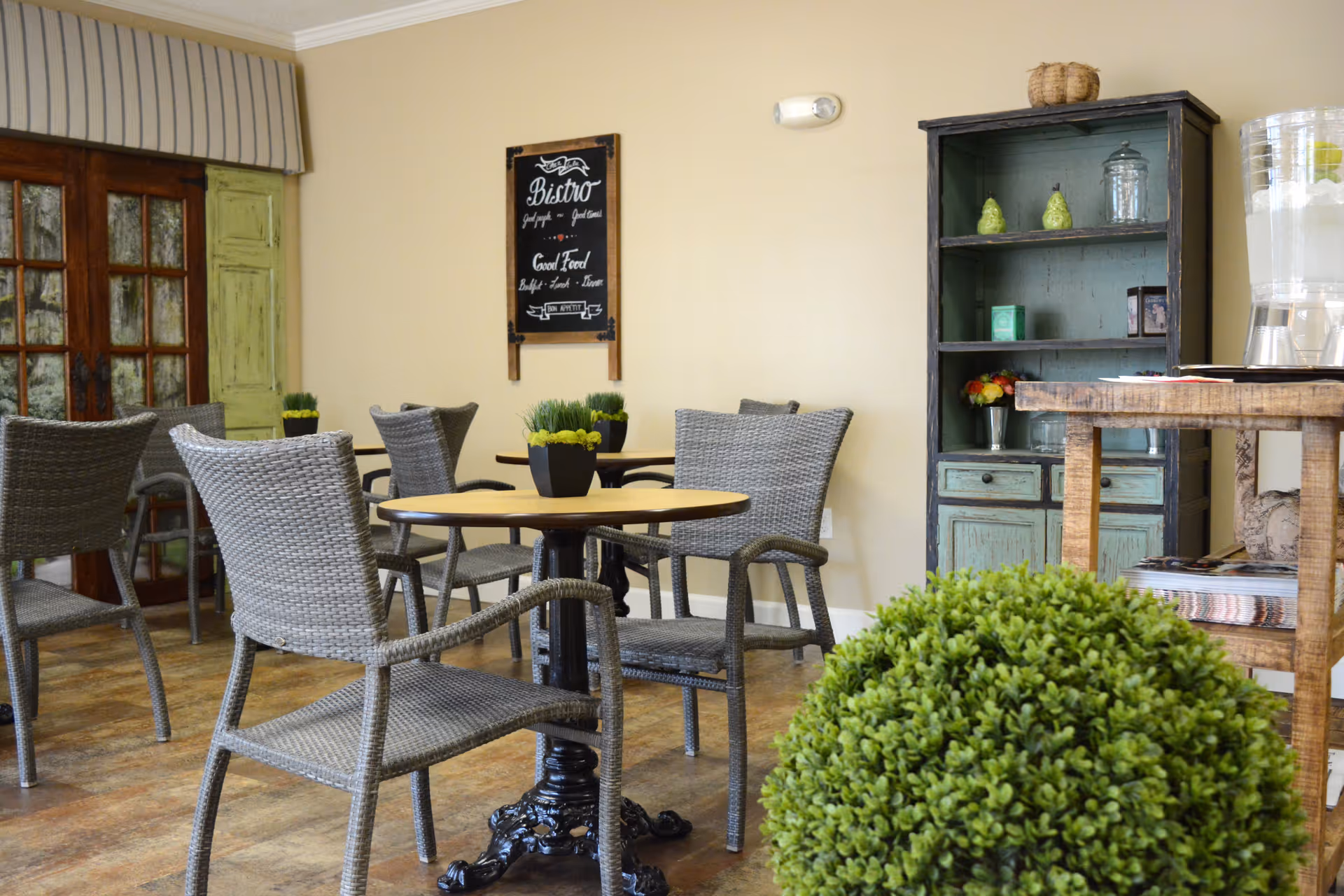Cozy dining area with wicker chairs around round tables, small potted plants, and a rustic shelving unit against a beige wall.