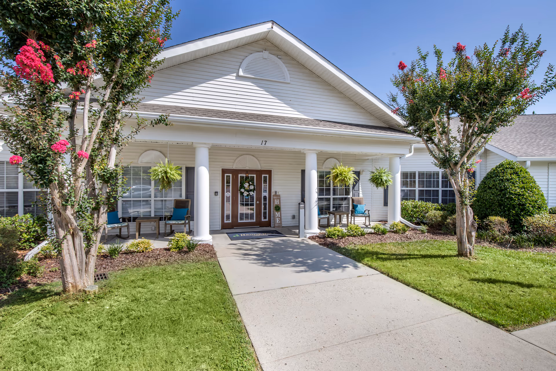 Front entrance of a single-story white building with a covered porch supported by four white columns. The porch has two seating areas with chairs and small tables, hanging green plants, and decorative elements including a wreath on the door and a sign spelling HOME. There are two flowering trees with pink blossoms on either side of the concrete walkway leading to the entrance, surrounded by green grass and shrubs under a clear blue sky.