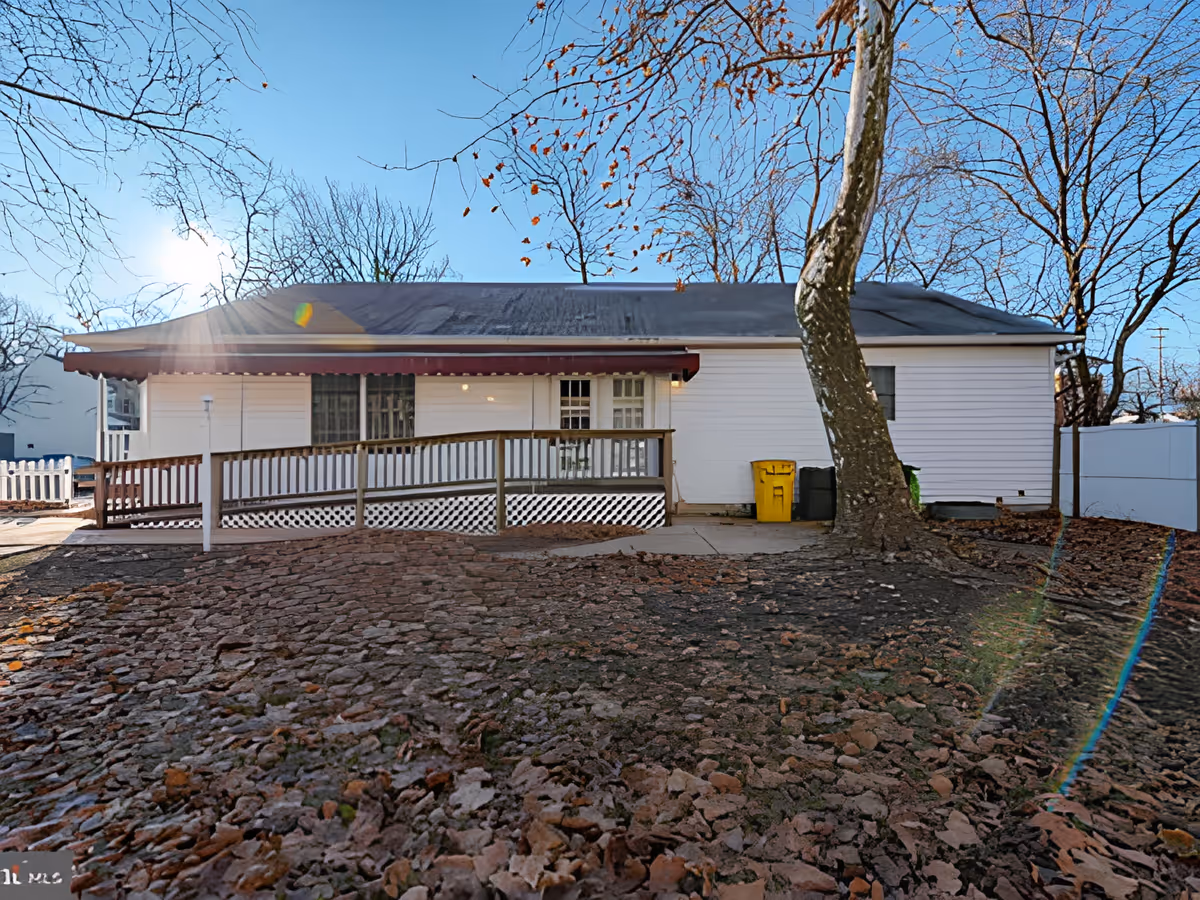 Exterior view of a single-story white building with a sloped roof, a wooden ramp leading to the entrance, a large tree in the foreground, and leaf-covered ground under a clear blue sky.