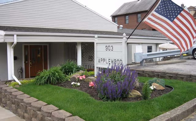 Front exterior view of a single-story building with a covered entrance, a garden bed with purple and pink flowers, a white sign displaying the address '903' and the name 'Applewood', and an American flag mounted on a pole near the sign.