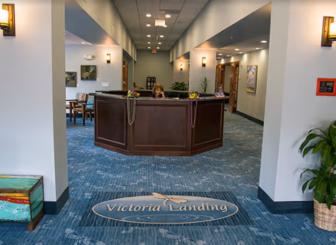 Reception area of Victoria Landing with a wooden front desk where a woman is seated. The floor has a blue carpet with the Victoria Landing logo. The walls are light-colored with modern wall sconces, and there are plants and framed artwork in the background.