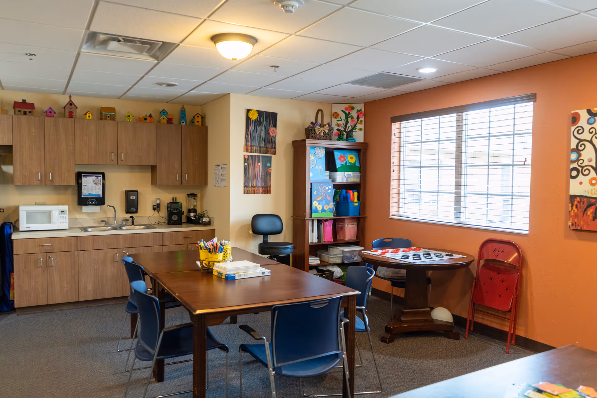 A bright room with a kitchenette featuring wooden cabinets, a microwave, a sink, and a coffee maker. There are two tables with blue chairs around them, one table has art supplies and books, and the other has a checkers board. A window with blinds lets in natural light, and colorful artwork and birdhouse decorations adorn the walls and shelves.