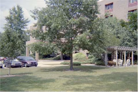 Exterior view of Oakbrook Towers with trees, cars, and a shaded outdoor seating area.