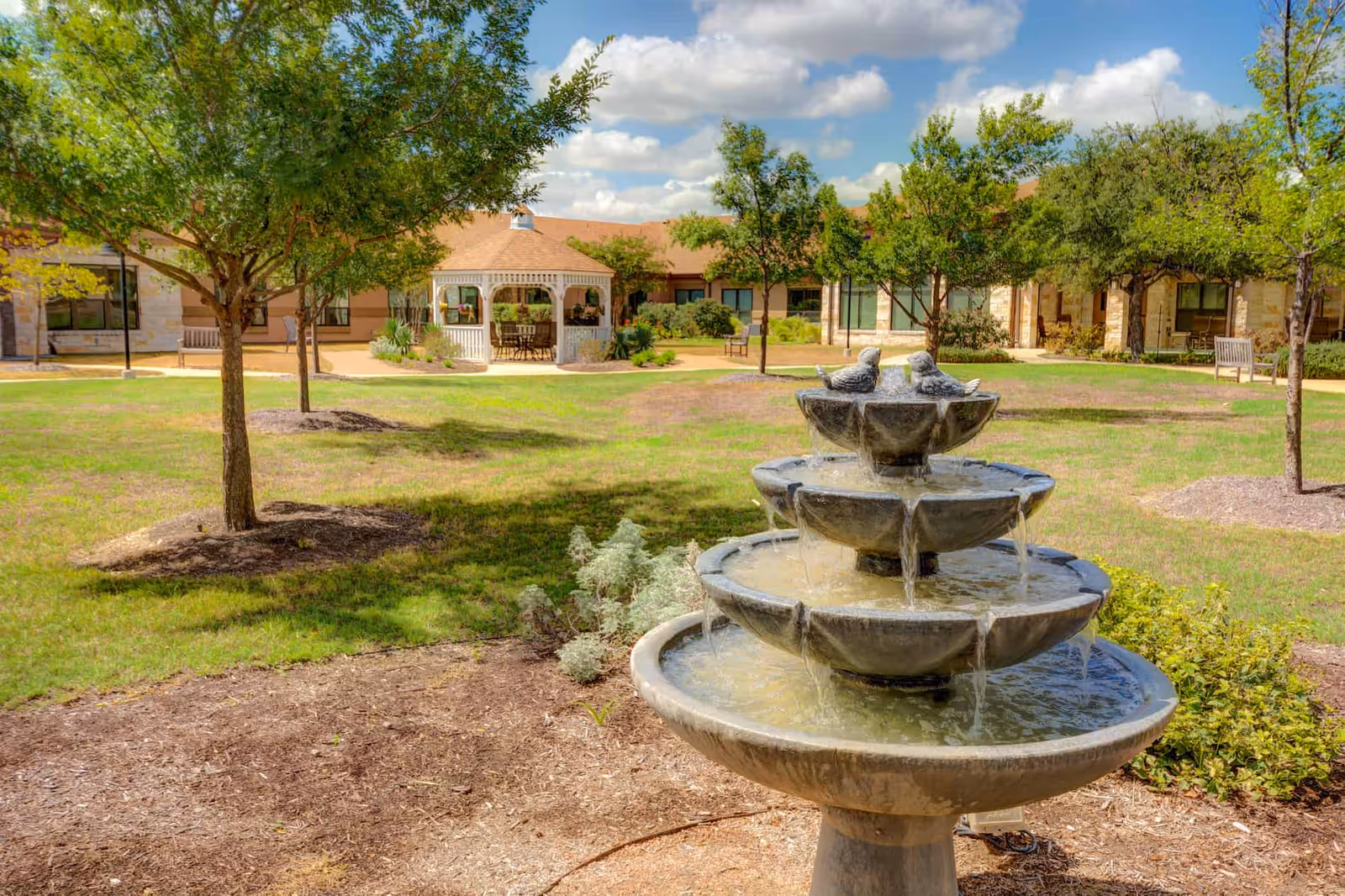 A multi-tier stone fountain in a grassy courtyard with trees, benches, and a gazebo in front of a single-story senior living building.