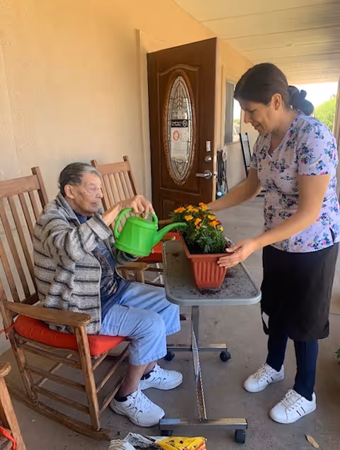 An elderly man sitting in a wooden rocking chair on a porch watering flowers in a rectangular planter while a woman stands next to him holding the planter. The porch has a wooden door with a decorative glass panel and beige walls.