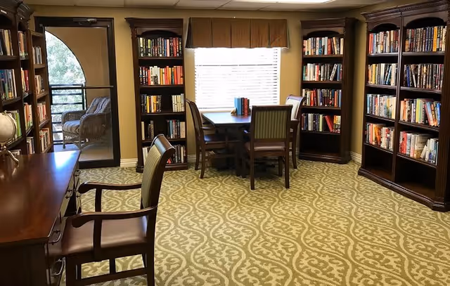 A cozy library room with patterned carpet, wooden bookshelves filled with books, a wooden table with four chairs near a window with blinds, and a wooden desk with a chair. A glass door leads to an outdoor seating area with wicker chairs.