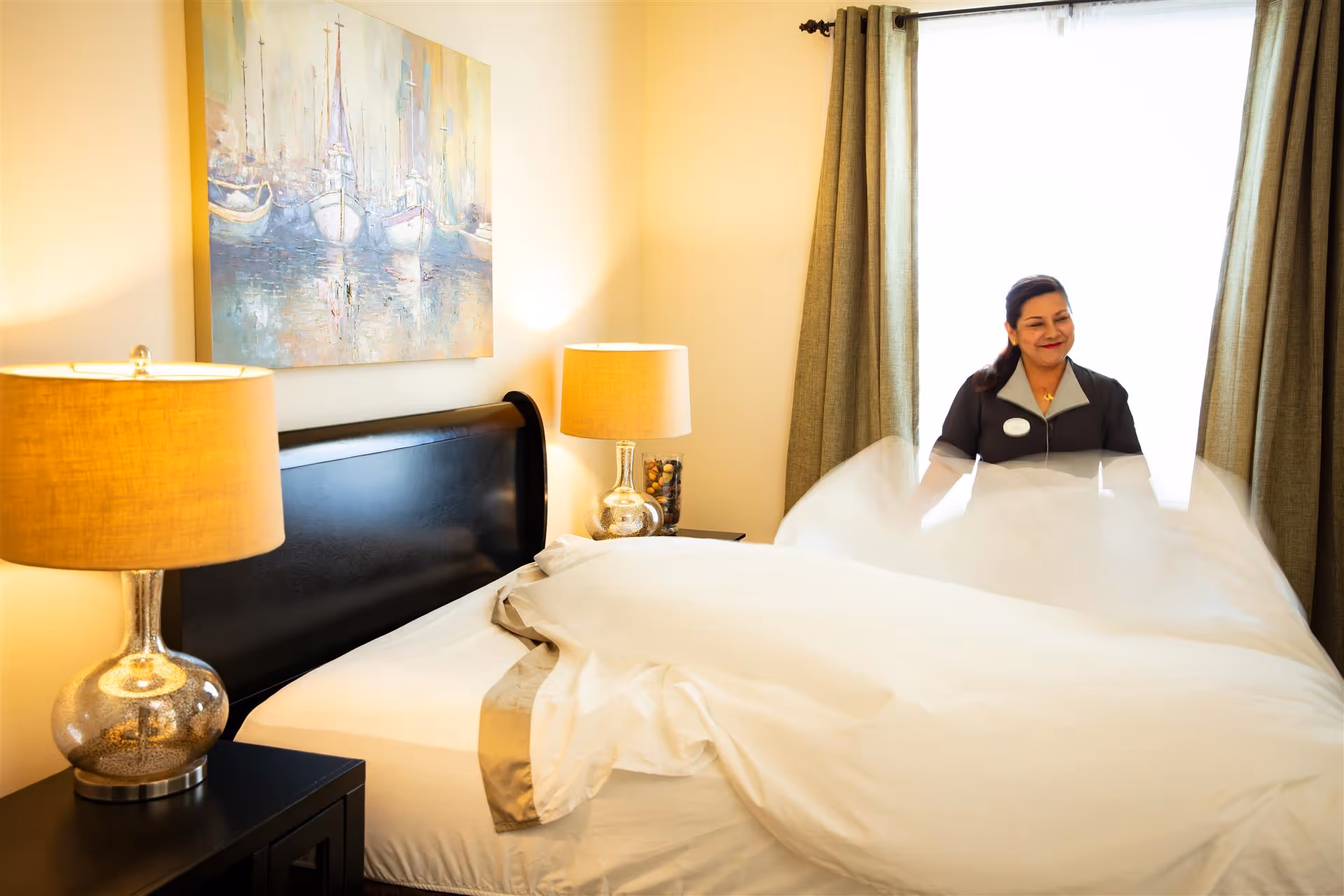 A woman is making a bed in a well-lit bedroom with two bedside lamps and a painting of boats on the wall above the headboard. The room has beige walls and curtains covering a window.