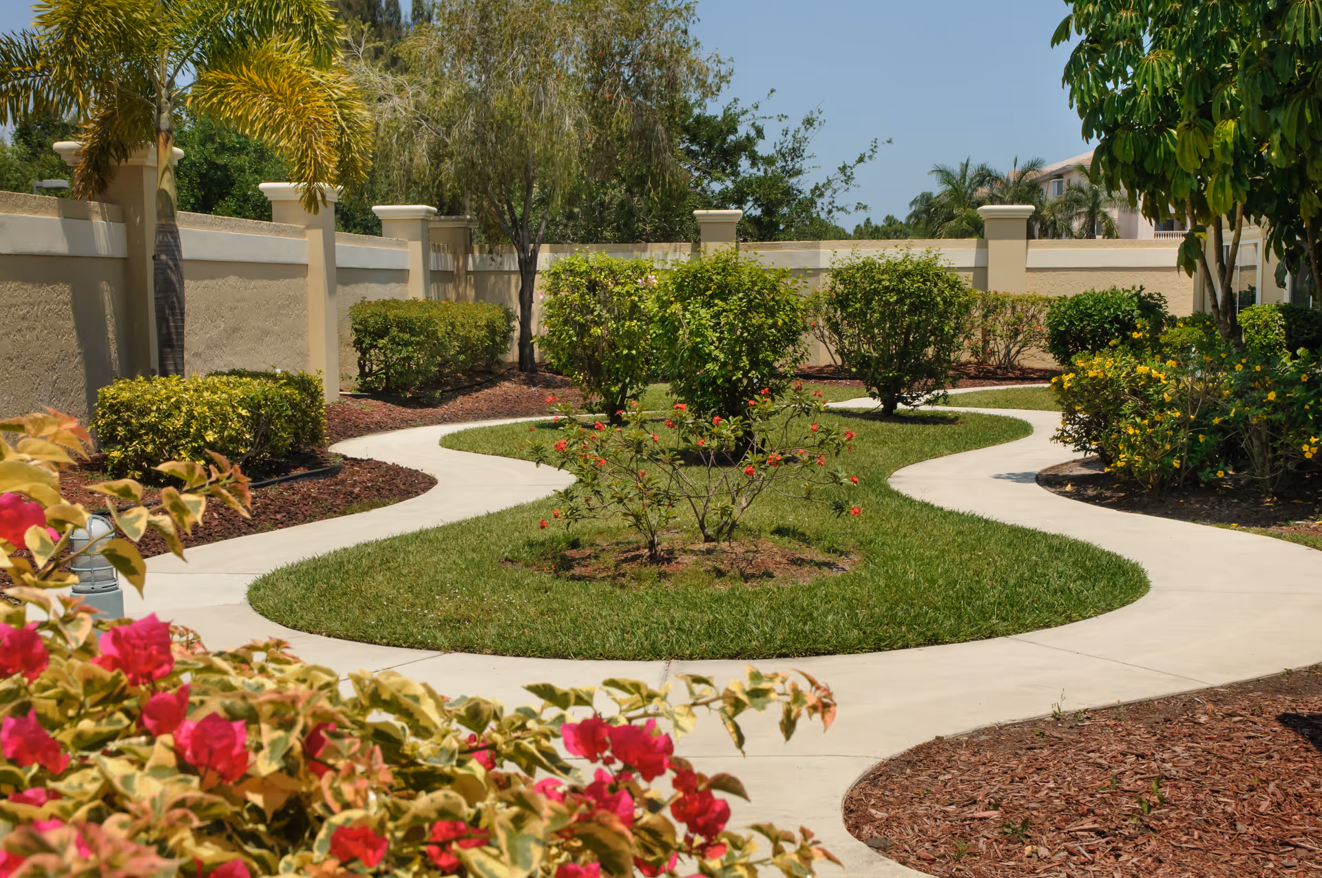 Curved concrete pathways winding through a landscaped garden area with green grass, various bushes, flowering plants, and trees, enclosed by a beige wall under a clear blue sky.