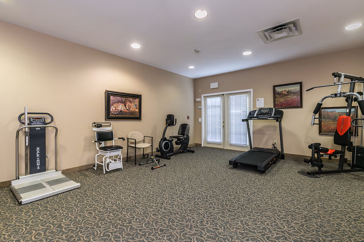 A fitness room with various exercise equipment including a scale, a chair with armrests, a recumbent exercise bike, a treadmill, and a multi-function weight machine. The room has beige walls, patterned carpet, recessed ceiling lights, and framed artwork on the walls. There are double glass doors with blinds.