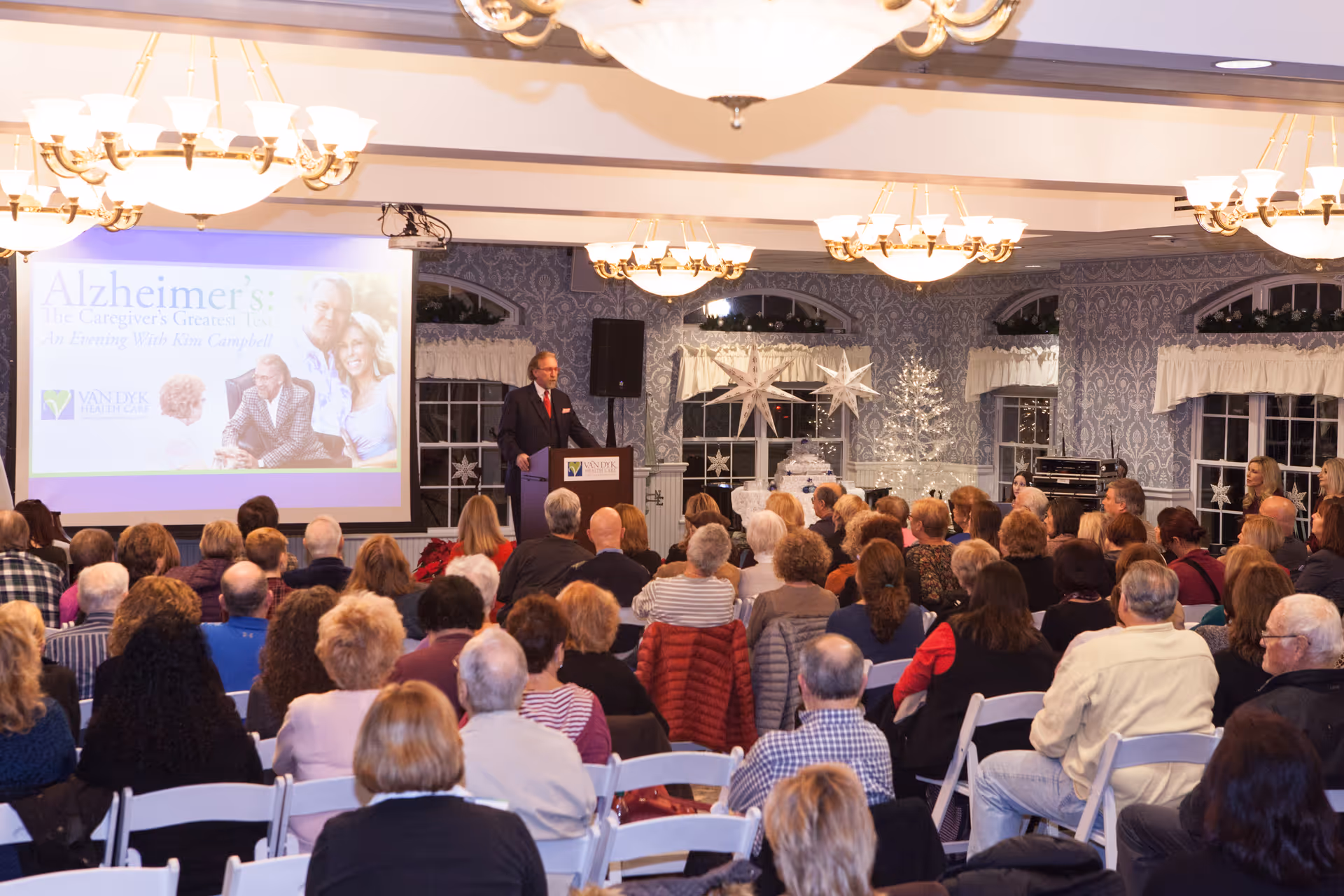 A large group of people seated in a decorated room attending a presentation about Alzheimer's titled 'Alzheimer's: the Caregiver's Greatest Test, An Evening With Kim Campbell.' A man in a suit stands at a podium speaking to the audience. The room has patterned wallpaper, large windows with white curtains, and festive holiday decorations including a white Christmas tree and star ornaments.