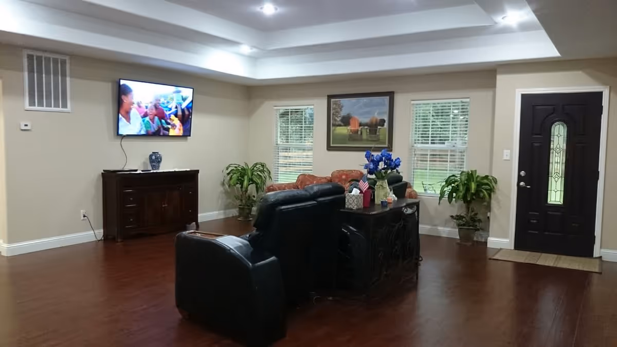 A cozy living room in a senior living facility with two black leather recliners and a patterned sofa arranged around a dark wooden console table. The room has hardwood floors, two windows with white blinds, a dark front door with decorative glass, and a wall-mounted TV showing a group of people. There are also two potted plants and a framed picture of outdoor chairs on the wall.