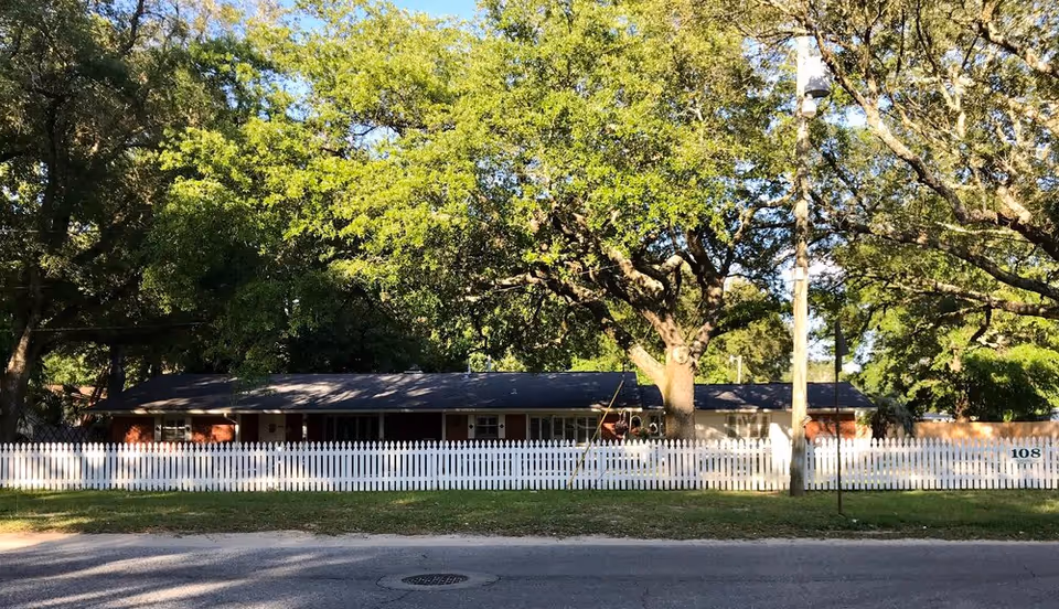 A single-story brick building with a dark roof behind a white picket fence, surrounded by large green trees and a grassy lawn, viewed from across the street on a sunny day.