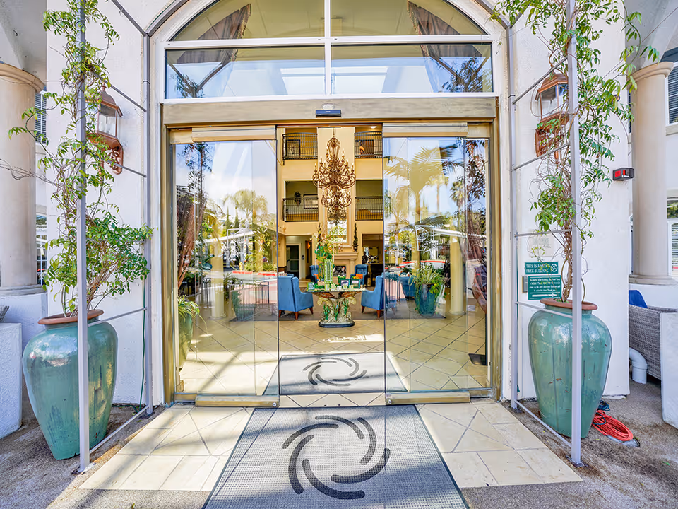 Glass automatic entrance doors to a building with large potted plants outside and a lobby with seating and a chandelier visible through the glass.