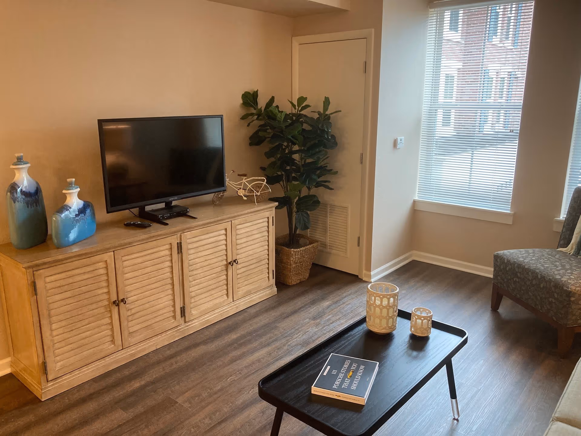 A cozy living room with a wooden TV stand holding a flat-screen television and two decorative blue vases. A small white decorative bicycle is also on the TV stand. To the right, there is a potted plant next to a closed door. In front, a black coffee table holds two candle holders and a book titled 'Power Stories That Leaders Should Know'. A patterned armchair is partially visible near two large windows with blinds, letting in natural light. The room has wood flooring and light-colored walls.