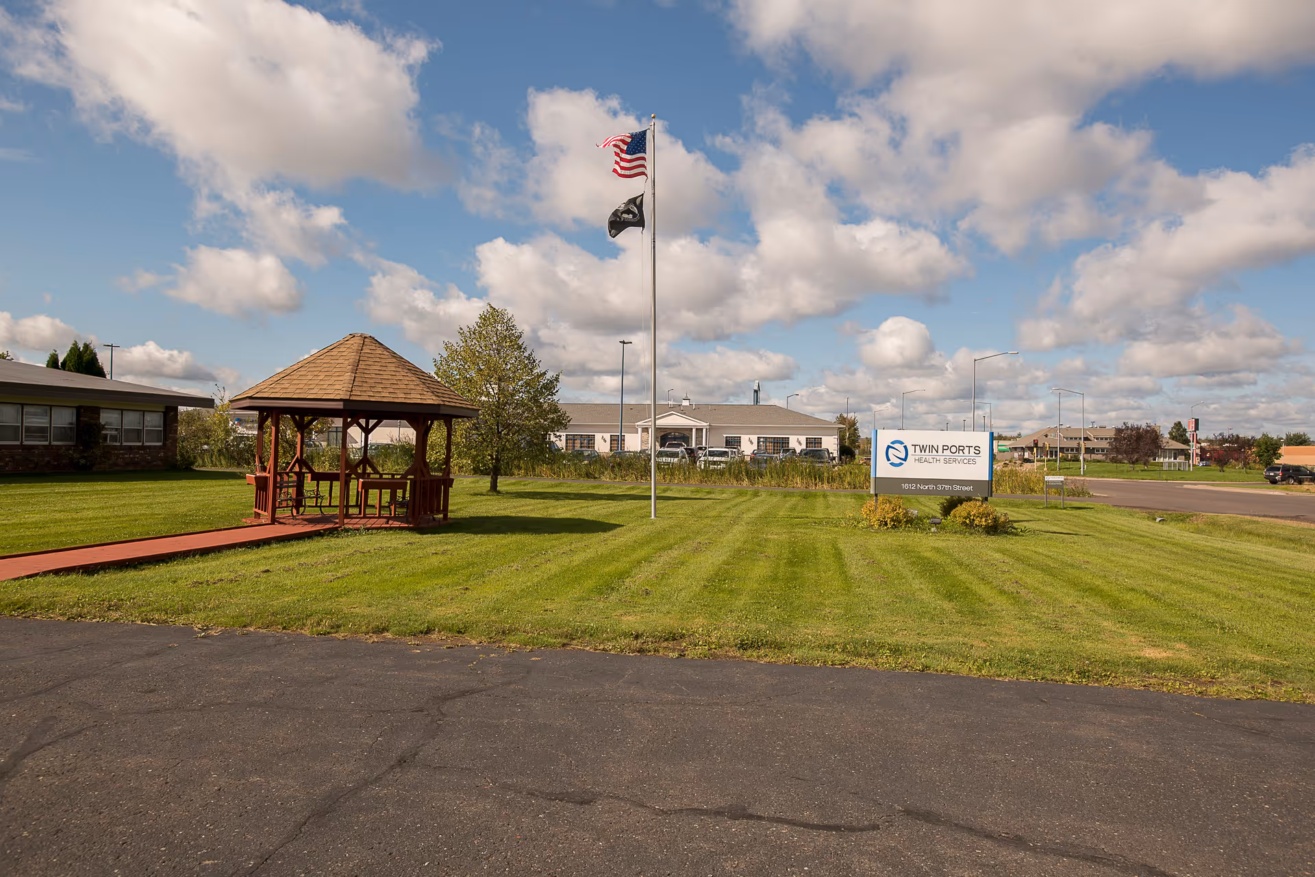 Outdoor view of Twin Ports Health Services facility with a green lawn, a wooden gazebo, two flagpoles with American and POW flags, and a sign displaying the facility name and address under a partly cloudy sky.