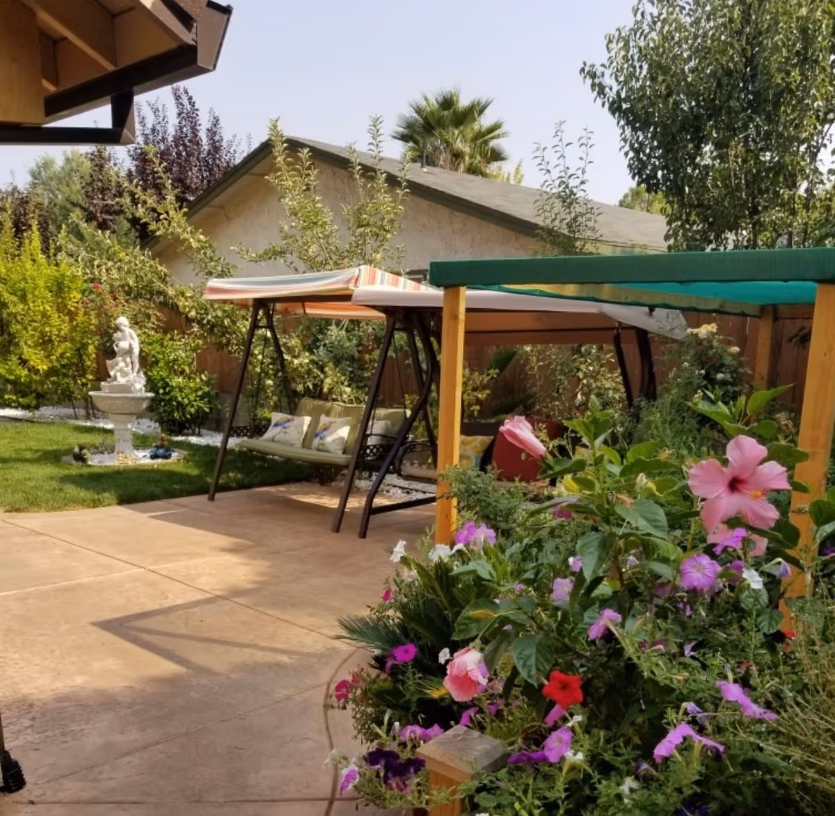 A peaceful outdoor garden area with a concrete patio, colorful flowers in the foreground, a cushioned swing with a striped canopy, green trees, and a small statue fountain in the background.