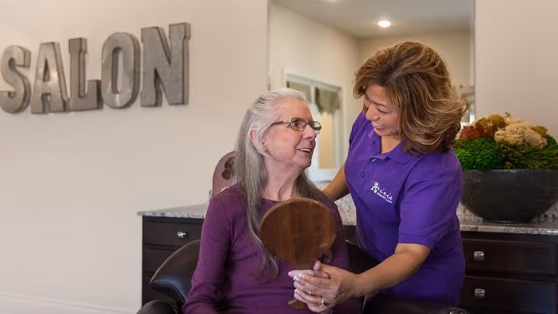 An elderly woman with long gray hair and glasses sits in a salon chair holding a hand mirror, while a smiling staff member in a purple shirt with the Iris Memory Care logo stands beside her, assisting and engaging with her. The word 'SALON' is displayed on the wall in large letters behind them.