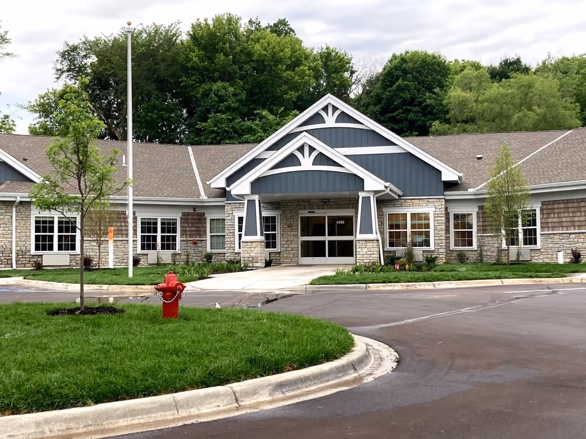 Front exterior view of NorBella Senior Living - Prior Lake building with a peaked roof entrance, stone and wood siding, surrounded by green grass, small trees, and a red fire hydrant in the foreground.