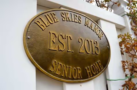 Bronze oval plaque on a white exterior wall reading Blue Skies Manor Est. 2013 Senior Home.