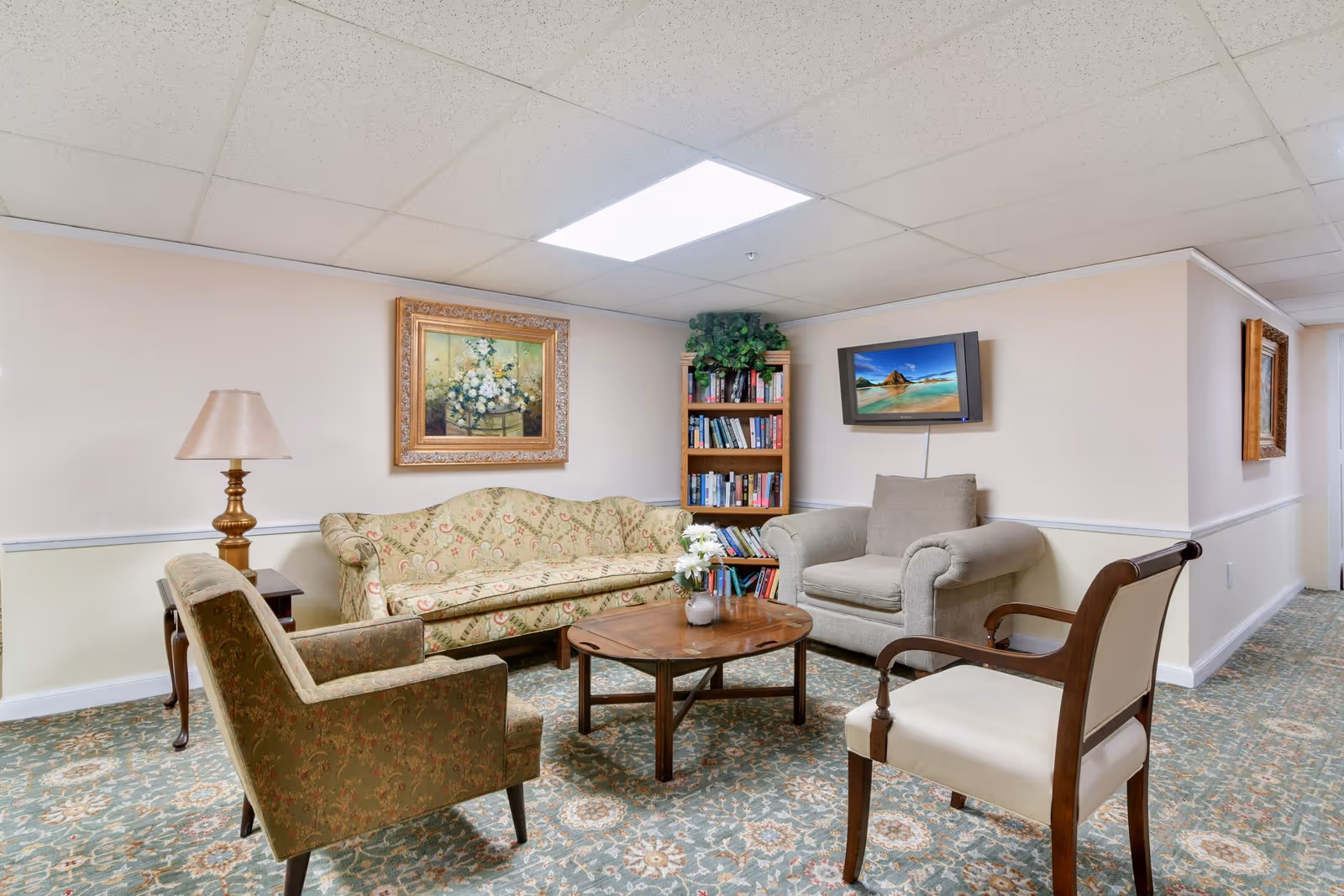 Cozy assisted-living lounge with sofas and armchairs arranged around a coffee table, a bookshelf, wall art and a mounted TV.