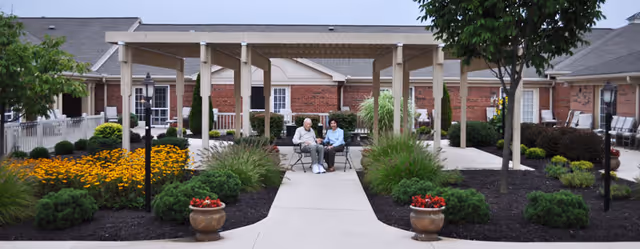 Two elderly people sit at a small table under a pergola in a landscaped courtyard in front of a brick senior living building.