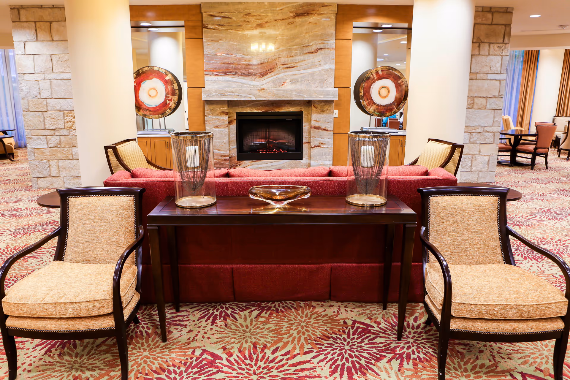 A cozy common room with a red sofa, two upholstered chairs, a wooden console table, and a fireplace.