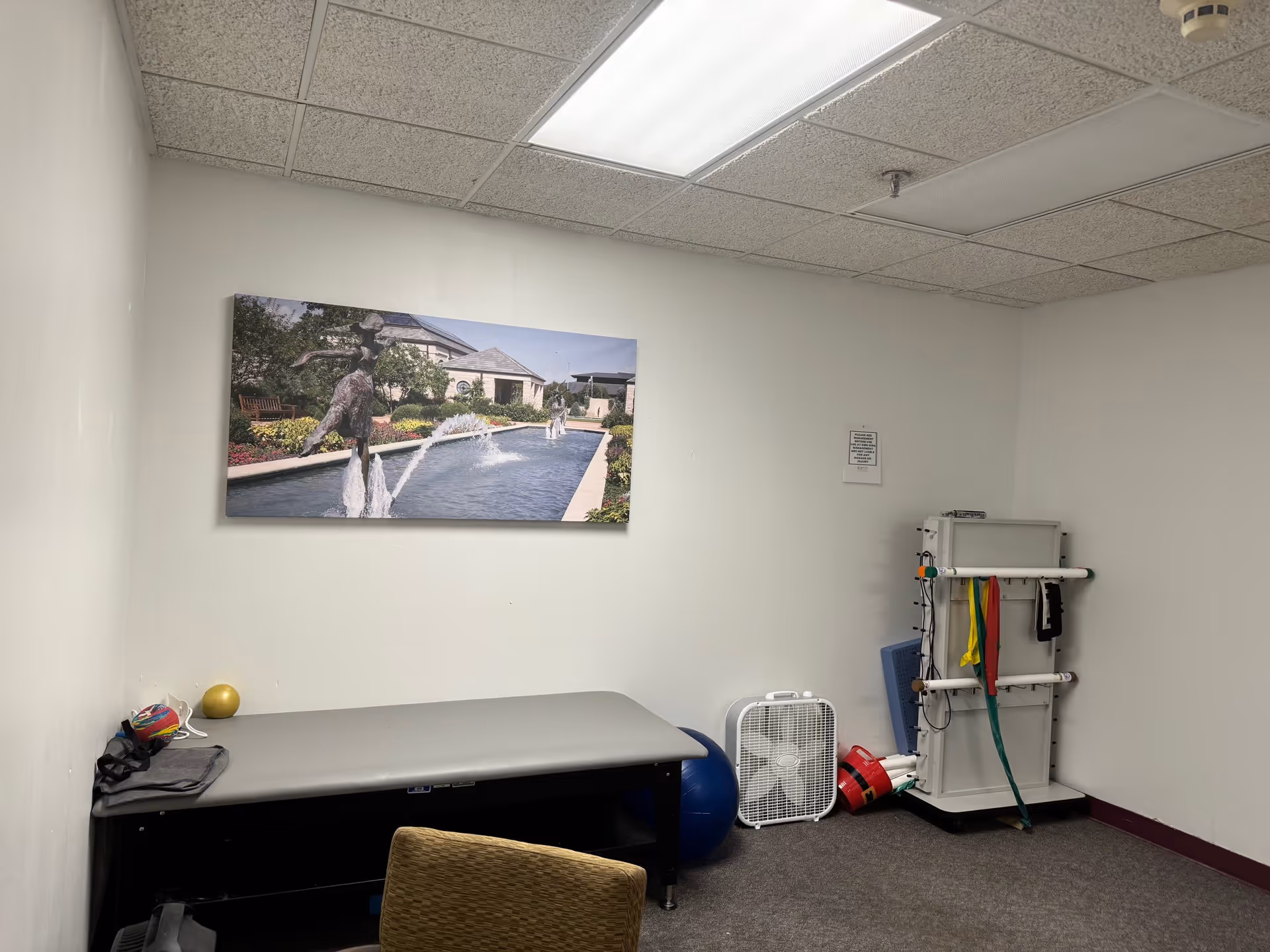 A small therapy or exercise room with a padded treatment table, exercise bands hanging on a rack, a blue exercise ball, a fan, and a chair. A large photo of an outdoor fountain with statues is mounted on the wall.