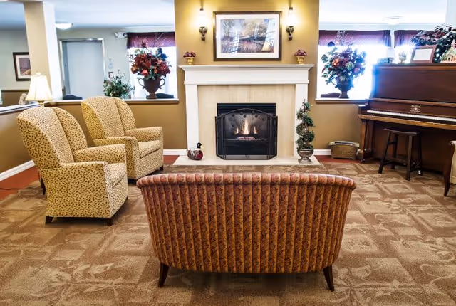 A cozy living room area with a lit fireplace centered on a beige wall. In front of the fireplace is a patterned sofa facing away from the camera, and to the left are two matching armchairs. On the right side, there is a wooden piano with a stool. The room is decorated with floral arrangements on either side of the fireplace and framed artwork above it. The carpet has a subtle floral pattern, and the walls are painted in warm tones.