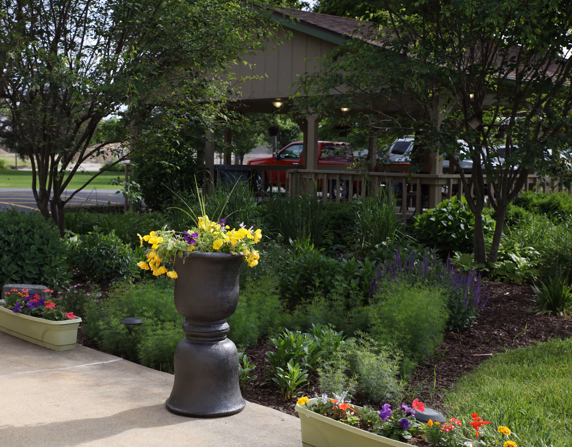 Outdoor garden area with various green plants and colorful flowers in planters and a large decorative urn. Trees and shrubs surround a covered pavilion with parked vehicles visible in the background.