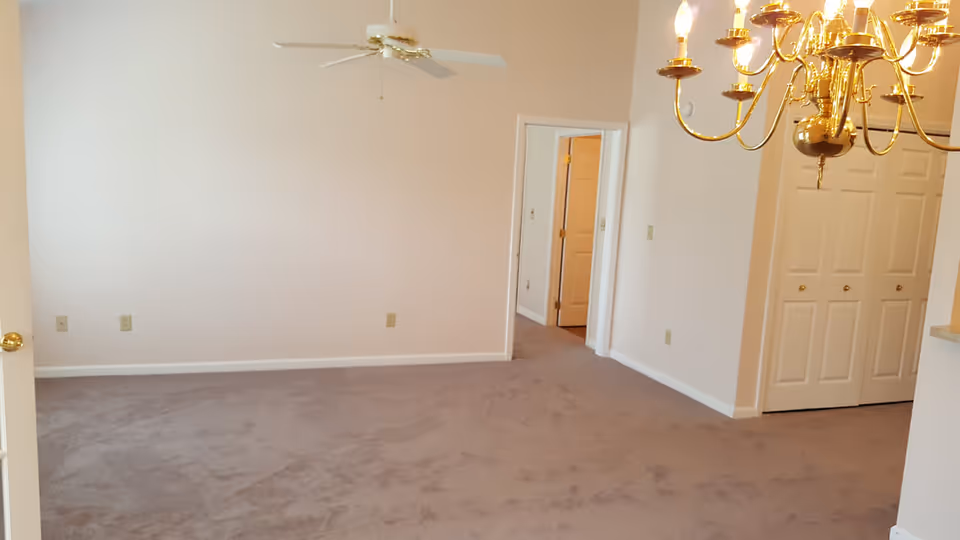 Empty carpeted living/dining area with a ceiling fan, brass chandelier, doorway, and double closet doors.
