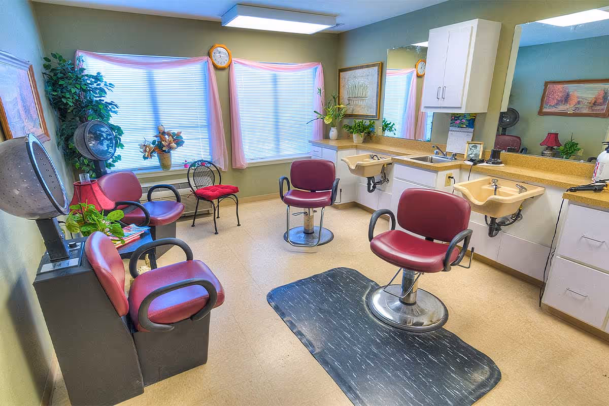 Interior of a hair salon area in a senior living facility with three red salon chairs, two beige sinks with mirrors above them, a hair dryer, and decorative plants and paintings near two windows with pink curtains.