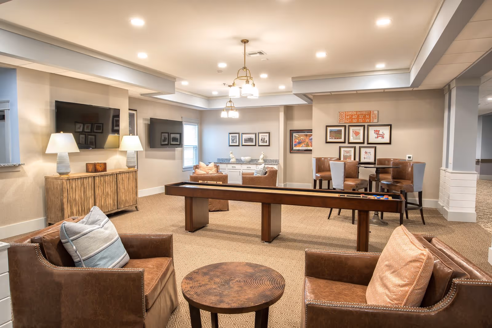 A spacious senior living common area featuring comfortable brown leather armchairs with cushions, a wooden shuffleboard table in the center, a round wooden side table, and a wooden cabinet with two table lamps. The walls are decorated with framed pictures and a sign. The room is well-lit with ceiling lights and has beige carpeting.