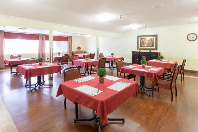 Dining room with several square tables draped in red tablecloths, each set with placemats and small potted centerpieces.