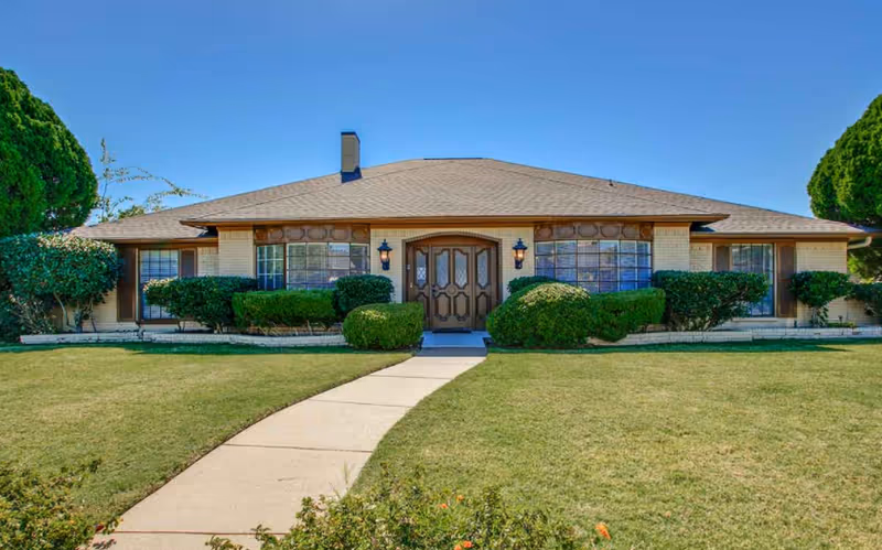Front exterior view of a single-story brick building with a tan roof, large windows, and a central double door entrance. The building is surrounded by neatly trimmed bushes and a well-maintained lawn with a concrete walkway leading to the entrance under a clear blue sky.