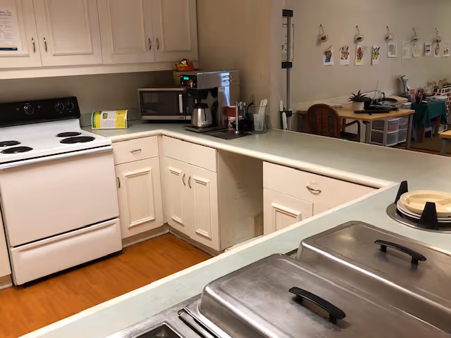 Institutional kitchen area with a stove, microwave, coffee maker, cabinets, countertops and stainless food warmers overlooking a seating area.