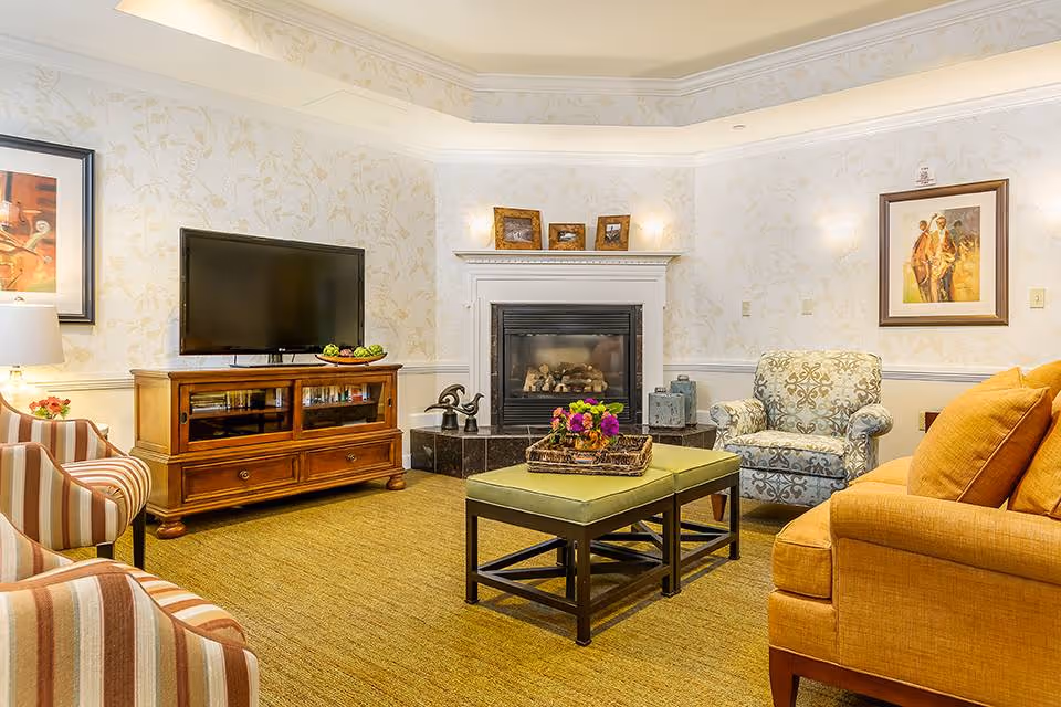 A cozy living room with a yellow sofa, a patterned armchair, and two striped armchairs. There is a wooden TV stand with a flat-screen TV on top, a fireplace with framed photos on the mantel, and a green ottoman coffee table with a basket of flowers. The walls have light floral wallpaper and framed artwork.