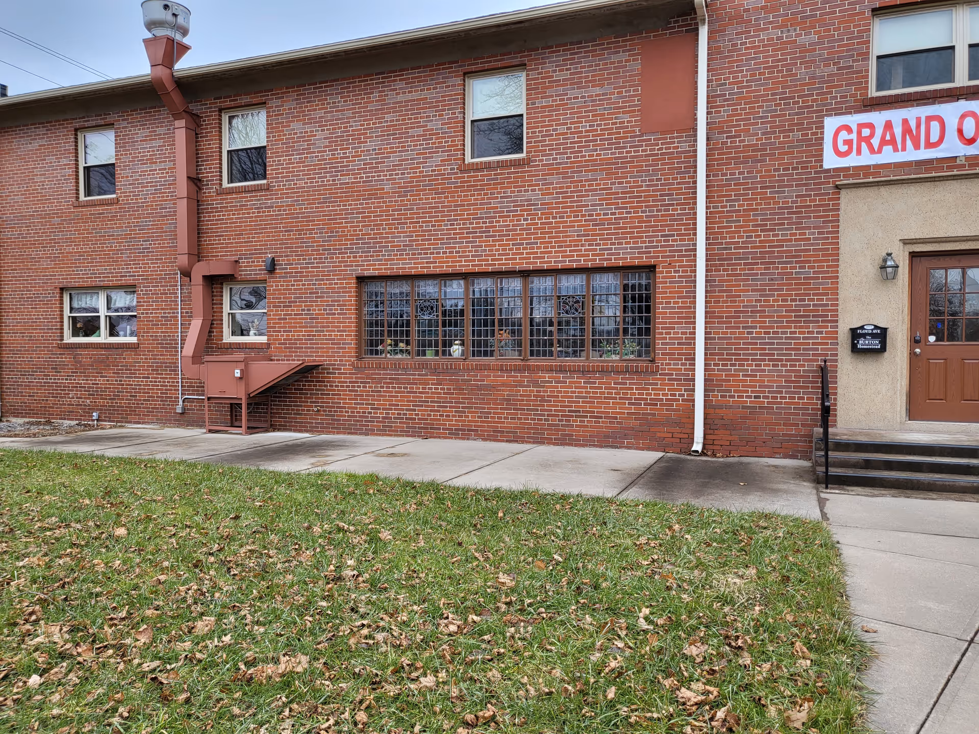Red brick building front with multi-pane windows, a brown entrance door and steps, a concrete walkway and a grassy lawn.