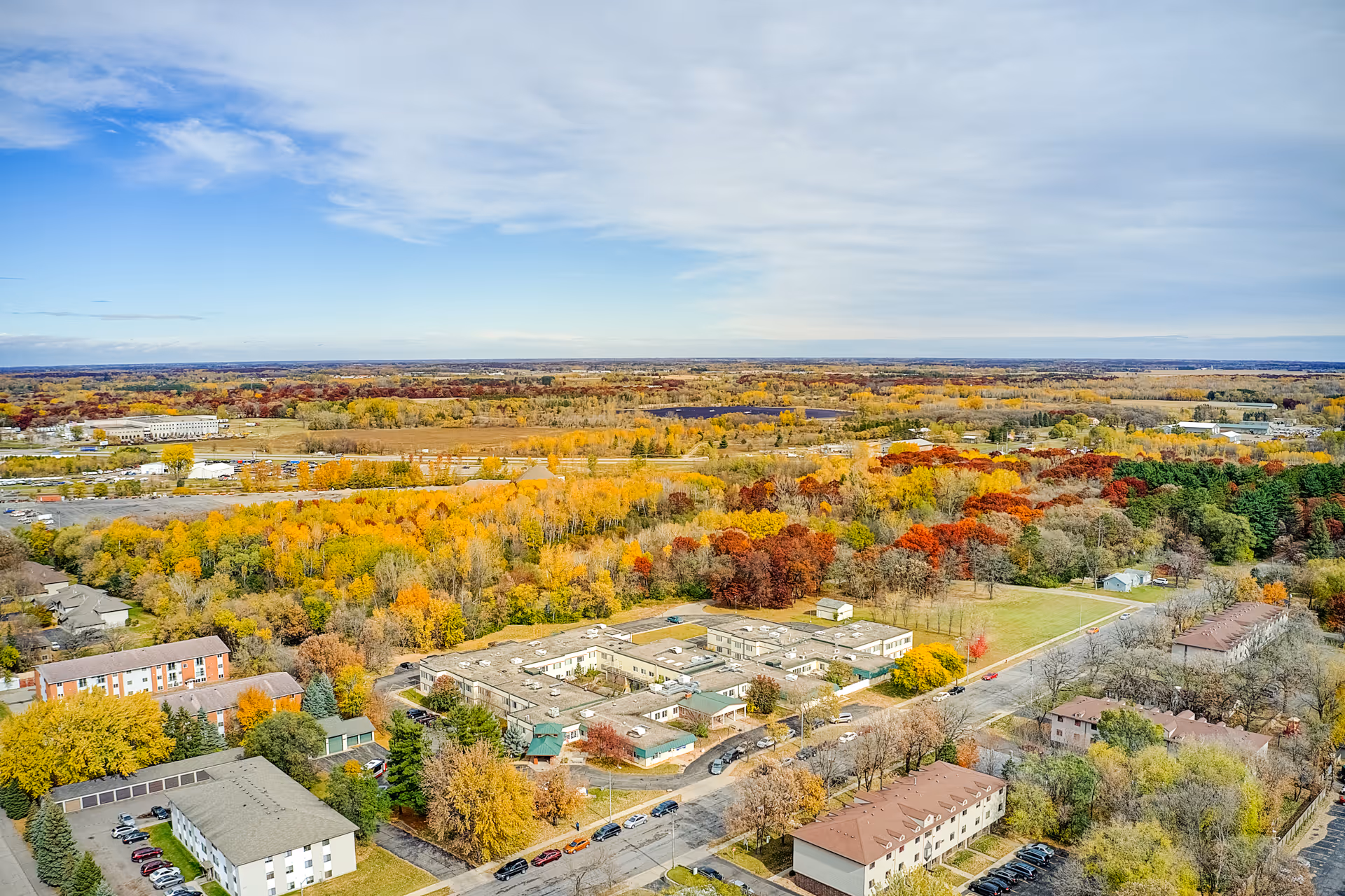 Aerial view of Nature's Point Assisted Living facility surrounded by trees with autumn foliage, nearby buildings, roads, and a vast landscape extending to the horizon under a partly cloudy sky.