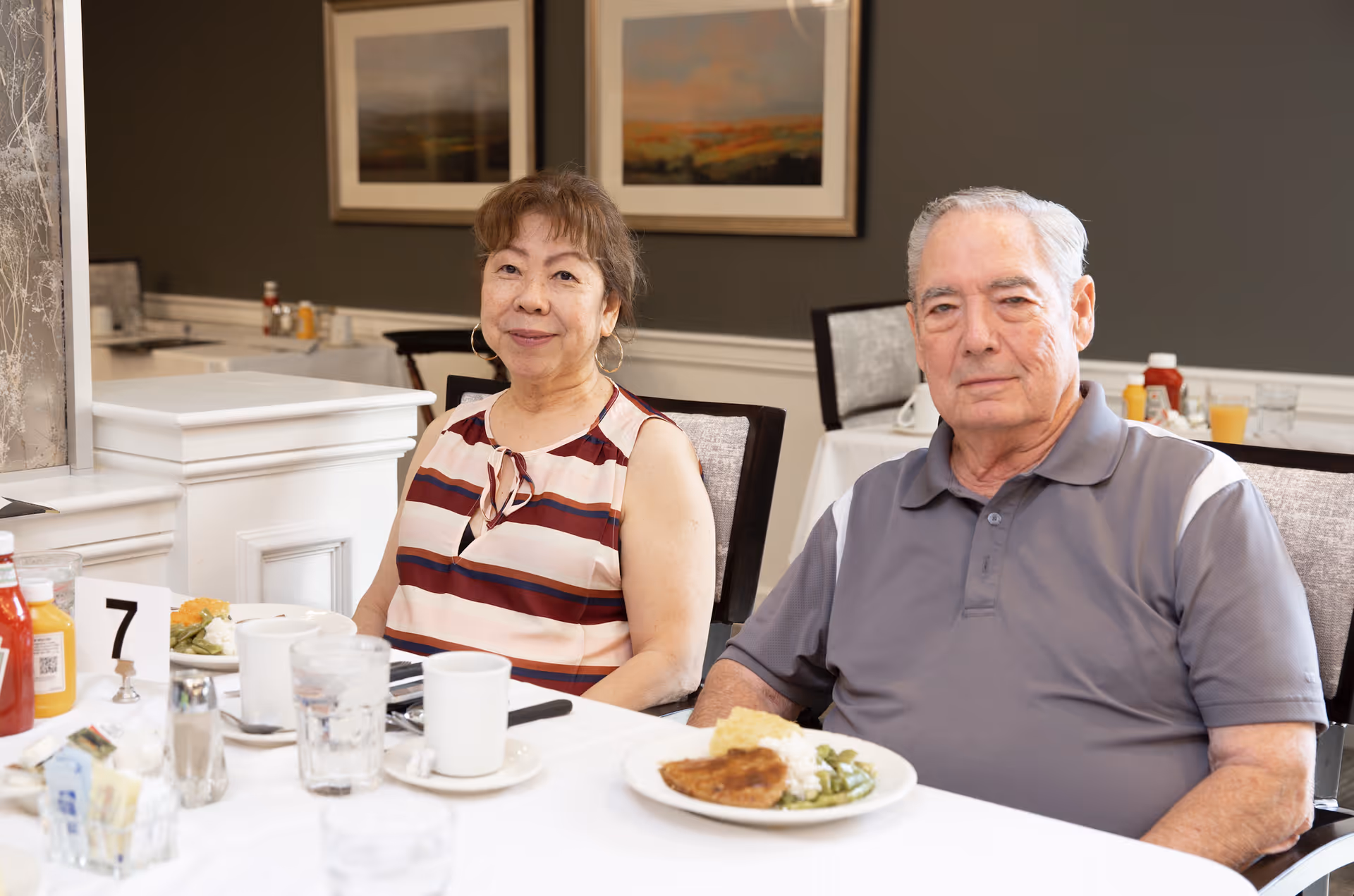 An elderly woman and man sitting at a dining table in a senior living facility. The woman is wearing a striped sleeveless top and the man is wearing a gray polo shirt. They have plates of food and cups in front of them. The background shows framed artwork on the wall and a neatly arranged dining area.