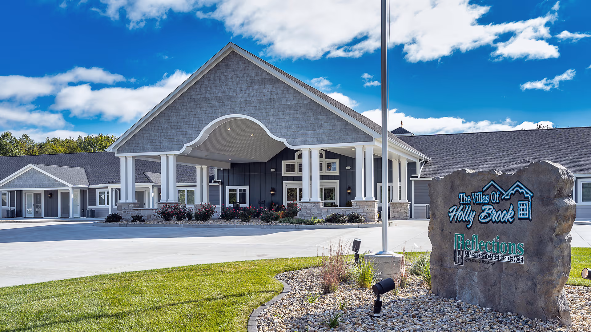 Front entrance of the Villas of Holly Brook assisted living and memory care building with a driveway and a stone sign in the foreground.