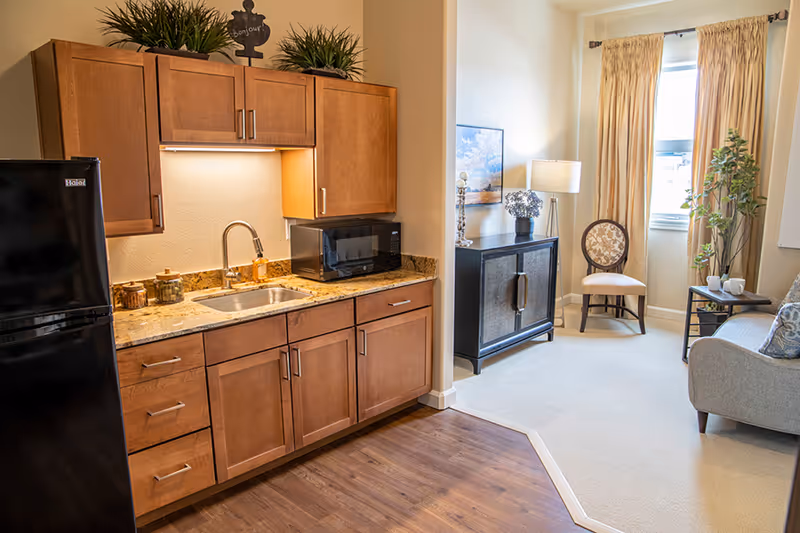 Bright senior living apartment interior with a kitchenette (wood cabinets, sink, microwave, fridge) opening to a furnished living area with a chair, cabinet, and window.
