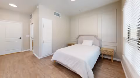 A bright bedroom with a neatly made bed featuring a white comforter and a single pillow. The room has light-colored walls with decorative molding, a wooden floor, two small wooden nightstands on either side of the bed, a closed white door, and a large window with white blinds allowing natural light to fill the space.