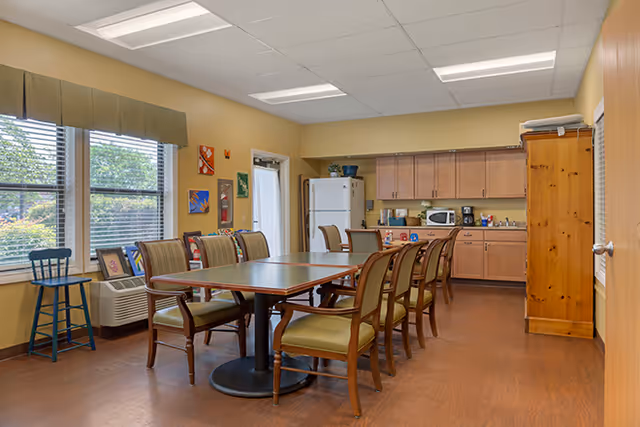 A dining room with a large rectangular table surrounded by eight wooden chairs with green cushions. The room has yellow walls, a window with a green valance, and several colorful paintings on the wall. In the background, there is a kitchenette area with wooden cabinets, a white refrigerator, a microwave, and a coffee maker. The floor is wooden, and the ceiling has fluorescent lighting.
