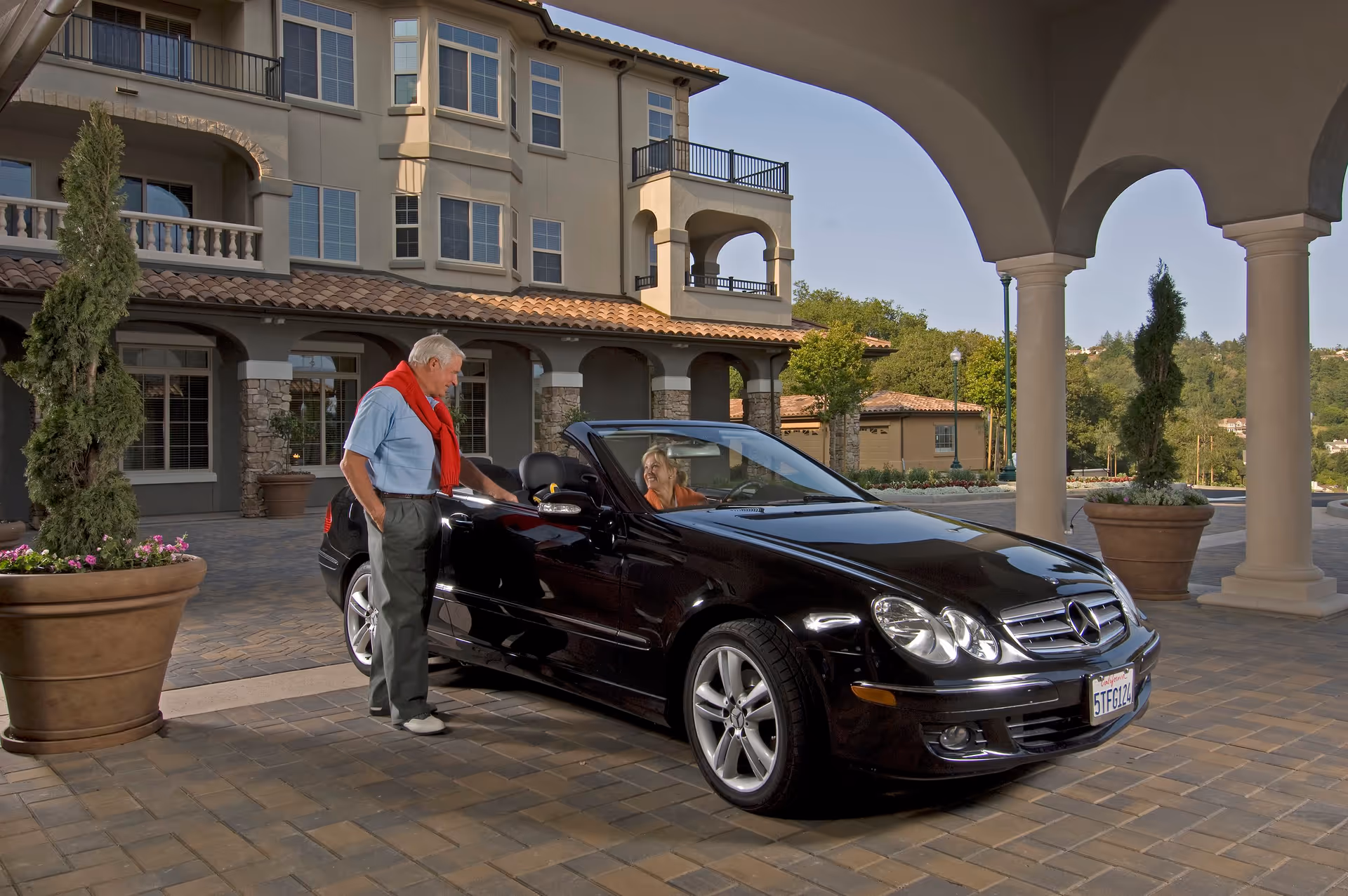 An elderly man with a red sweater draped over his shoulders stands next to a black Mercedes convertible car, talking to an elderly woman seated inside the car. They are outside a large, elegant building with arches, balconies, and potted plants, under a covered driveway.