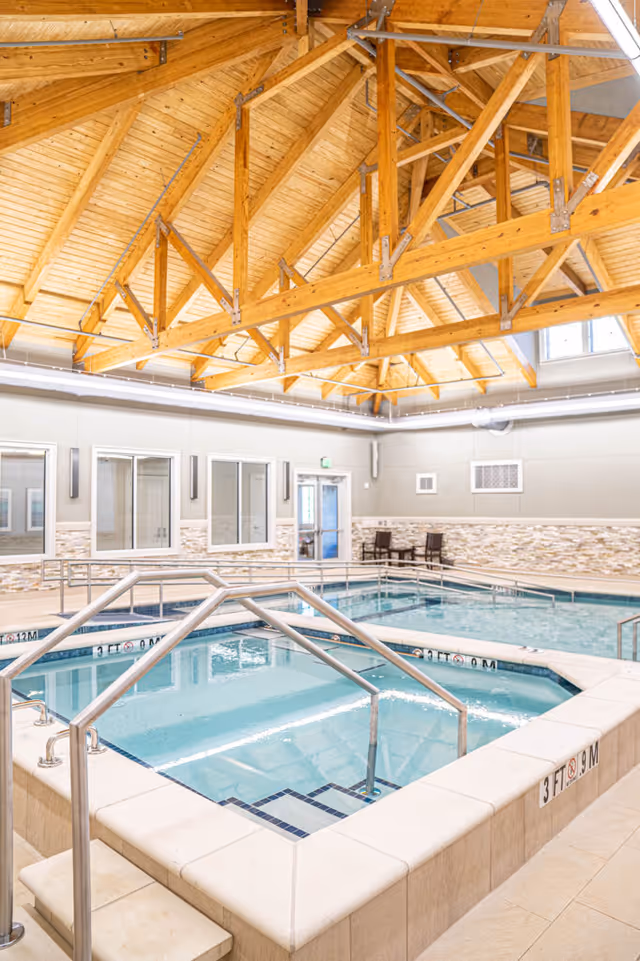 Indoor therapy pool and hot tub with stainless handrails beneath a vaulted wooden beam ceiling in a senior living facility.