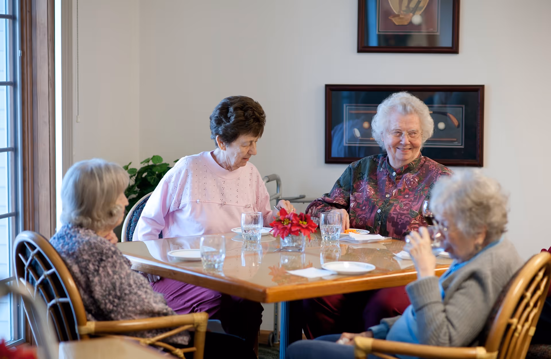 Four elderly women sitting around a wooden dining table with glasses and plates, engaging in conversation in a well-lit room with framed artwork on the wall and a window letting in natural light.
