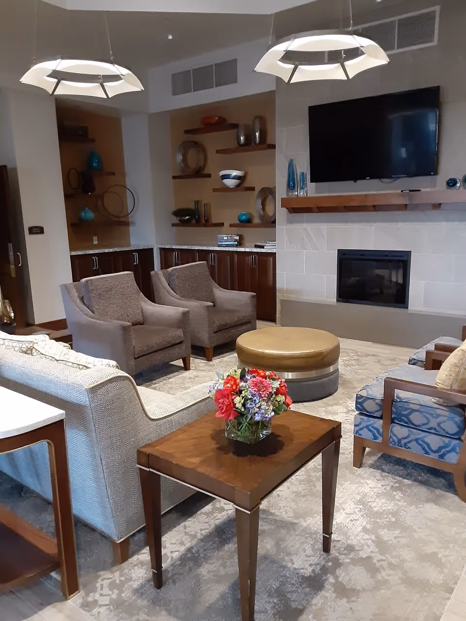 A cozy living room area with two gray armchairs, a beige sofa, and two blue patterned chairs arranged around a round ottoman. A wooden side table with a colorful flower arrangement sits in the foreground. The room features a modern fireplace with a mounted flat-screen TV above it, and decorative shelves with vases and bowls on the back wall. Two contemporary pendant lights hang from the ceiling.